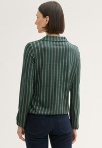 Langärmlige Bluse aus dunkelgrünem Stoff mit vertikalem weißen Streifenmuster und Kragen. Weiche Textur, taillierter Schnitt, kontrastierende Manschettendetails.