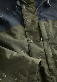 Jachetă rezistentă la apă, de culoare verde măsliniu și neagră, cu picături de apă pe țesătură, care arată mâneca și capsele din față.