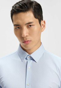 Camicetta azzurro chiaro con un sottile motivo geometrico, colletto button-down e tessuto testurizzato. Presenta bottoni standard nella parte anteriore.