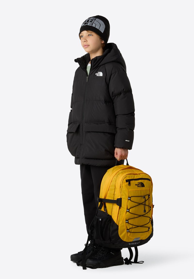 Chaqueta de plumas negra con capucha, mochila amarilla y negra con diseño de cordón elástico, y botas negras. Un fondo neutro mejora la visibilidad.