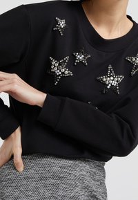 Felpa nera con applicazioni a forma di stella decorate con perle e gemme. Collo a costine e gonna grigia strutturata visibile in vita.