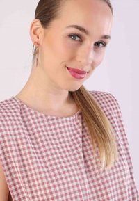 Ärmelloses Top mit Rosenmuster, präsentiert einen runden Ausschnitt und ein Karomuster in Rosa und Weiß. Das Model trägt große Creolen und hat lange Haare.