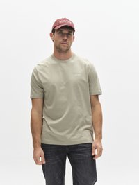 Olivgrön bomulls-t-shirt med korta ärmar och rund halsringning, kombinerad med mörka jeans och en vinröd keps med vit text.