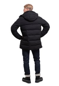 Chaqueta impermeable negra con capucha, con un diseño acolchado y puños elásticos. Llevada sobre jeans de mezclilla oscuros y calzado negro.