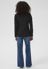 Blazer noir sur mesure à la coupe ajustée, avec fentes au dos, associé à un jean évasé à délavage clair arborant des coutures discrètes sur les poches.