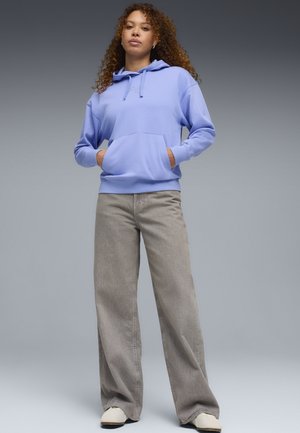 Vrouw staand met een lichtblauwe hoodie met handen in de voorzak, grijze wijde jeans en witte schoenen tegen een grijze achtergrond.