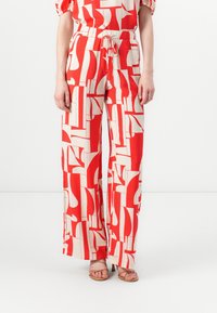 Pantalons larges à motifs rouges et crème, avec une taille élastique et des poches latérales, présentant des formes géométriques et une texture lisse.