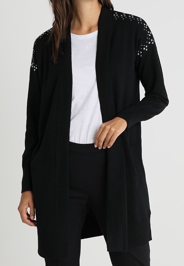 Femme portant un long cardigan noir avec des clous argentés aux épaules sur un t-shirt blanc et un pantalon noir, debout devant un fond uni.