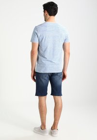 Man som står med ryggen mot kameran, iförd en ljusblå och vit randig t-shirt, mörka denimshorts och vita sneakers, mot en vit bakgrund.