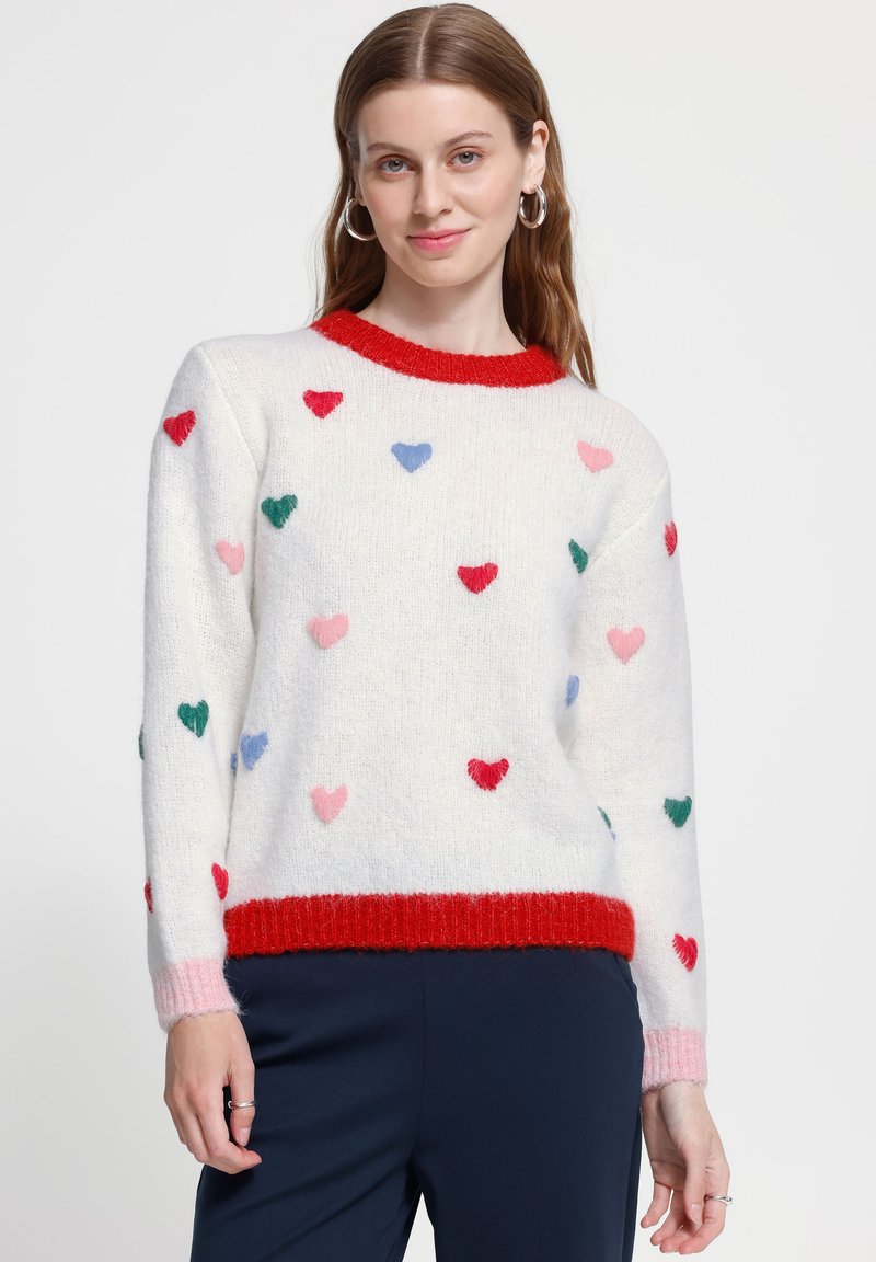 Femme portant un pull blanc avec des motifs de cœurs rouges, bleus, roses et verts, avec des bordures rouges et roses, associé à un pantalon bleu foncé.