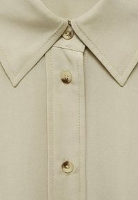 Light beige collared shirt featuring a front button placket with round beige buttons, smooth fabric texture, and contrasting stitching detail.