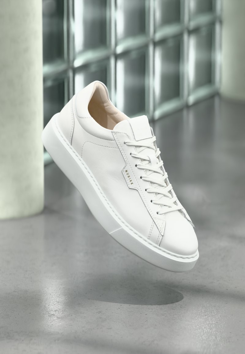 White leather sneakers with a rounded toe, lace-up design, and subtle branding. Features a smooth texture and a contrasting white rubber sole.