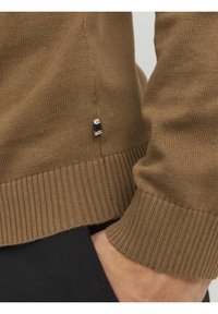 Brown knitted sweater with ribbed hem and cuffs, featuring a small, branded tag in contrasting colors near the side seam.