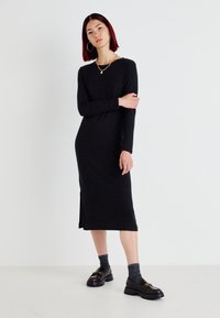 Black, long-sleeve ribbed dress with side slit, paired with dark socks and chunky black shoes featuring gold hardware accents.