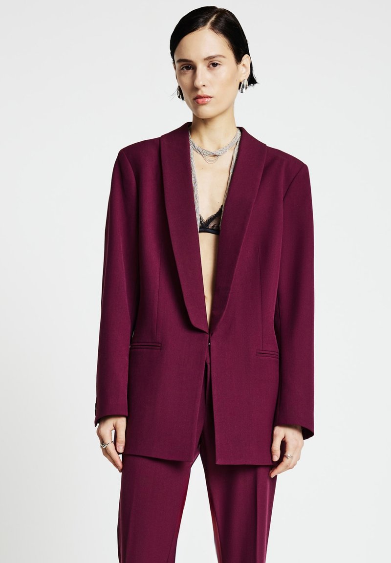 Bordeauxroter, maßgeschneiderter Blazer mit tiefem V-Ausschnitt, strukturierten Schultern und zwei Vorderentaschen, kombiniert mit passenden Hosen.