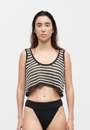 Νεαρή γυναίκα με μακριά σκούρα μαλλιά που φοράει μαύρο-άσπρο ριγέ πλεκτό crop top πάνω από μπικίνι τοπ και μαύρα μπικίνι κάτω, στέκεται ακίνητη.
