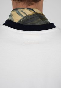 Vue arrière d'une personne portant une chemise blanche avec un col orné de noir et une écharpe beige à motifs autour du cou.