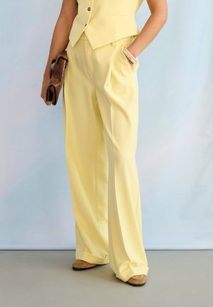 Naine, kes kannab kahvatukollaseid laiade säärtega pükse ja vest, hoides pruuni käekotti, üks käsi taskus ning nähtaval beežid kingad.