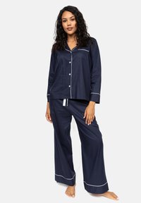 Navyblauwe pyjama set met lange mouwen en witte piping details. De top heeft een zak en knopen; de broek heeft een wijde pijp en een elastische trekkoord.