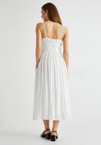 Witte midi-jurk met verstelbare spaghettibandjes, gerimpelde lijfje en een uitlopende rok. Gecombineerd met zwarte sandalen met chunky hakken.