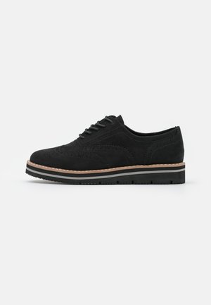 Black suede wingtip oxford shoe with lace-up front, beige trim on the sole, and black ridged platform sole on white background.