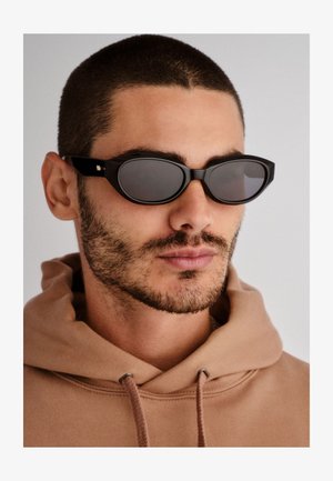 Fekete napszemüveg ovális lencseformával, sötétített lencsékkel és vastag kerettel. Diszkrét arany kiegészítők a szárakon.