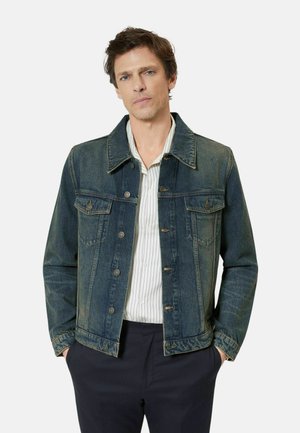 Mężczyzna w ciemnoniebieskiej kurtce dżinsowej na białej koszulce w paski i czarnych spodniach, stojący z rękami w kieszeniach na jednolitym tle.