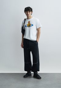 Λευκό κοντομάνικο μπλουζάκι με πολύχρωμο γραφικό σχέδιο, συνδυασμένο με μαύρο παντελόνι με φαρδιά μάνικα. Φορεμένο με μαύρη τσάντα ώμου και αθλητικά παπούτσια.