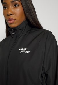 Jaqueta preto com fecho éclair, gola alta e textura suave. Apresenta logo bordado em branco no peito. Design leve e ajustado.