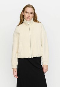 Cream bomber jacket with a high collar, textured fabric, zip closure, and side pockets. Worn over a black skirt. Smooth, lightweight material.