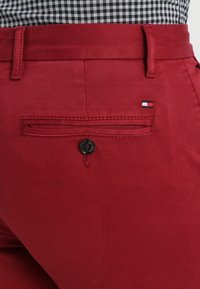 Nahaufnahme der Gesäßtasche einer roten Hose mit Knopf, Gürtelschlaufen und einem kleinen rot-weiß-blauem Flaggenlogo auf dem Stoff nahe der Tasche.