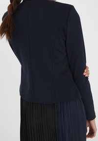 Tmavě modré sako na míru s dlouhými rukávy, hladkou texturou a střiženou siluetou, zobrazené zezadu přes plisovanou sukni.