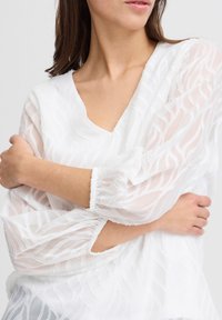Weißes Blusenshirt mit V-Ausschnitt, transparentem Stoff und strukturiertem Wellenmuster. Mit langen, gepufften Ärmeln und elastischen Bündchen sowie einer lockeren Passform.