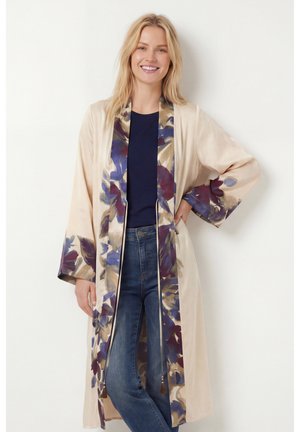 Femme souriante portant un kimono long beige à fleurs sur un haut bleu marine et un jean bleu, debout avec une main sur la hanche.
