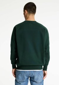 Hombre de espaldas vistiendo una sudadera verde oscura y jeans azules, con las manos relajadas a los lados sobre un fondo blanco.