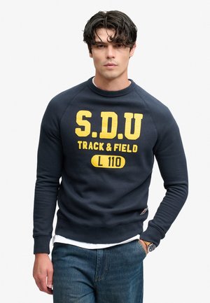 Młody mężczyzna o ciemnych kręconych włosach, ubrany w granatową bluzę z żółtym napisem "S.D.U Track & Field L 110" i niebieskie dżinsy, stoi z jedną ręką w kieszeni.