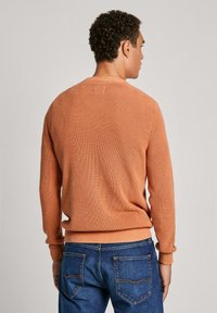 Rebrast pulover v oranžni barvi z dolgimi rokavi in okroglim izrezom, v kombinaciji z modrimi kavbojskimi hlačami. Strukturirano pleteno blago, oprijeto v pasu in na manšetah.