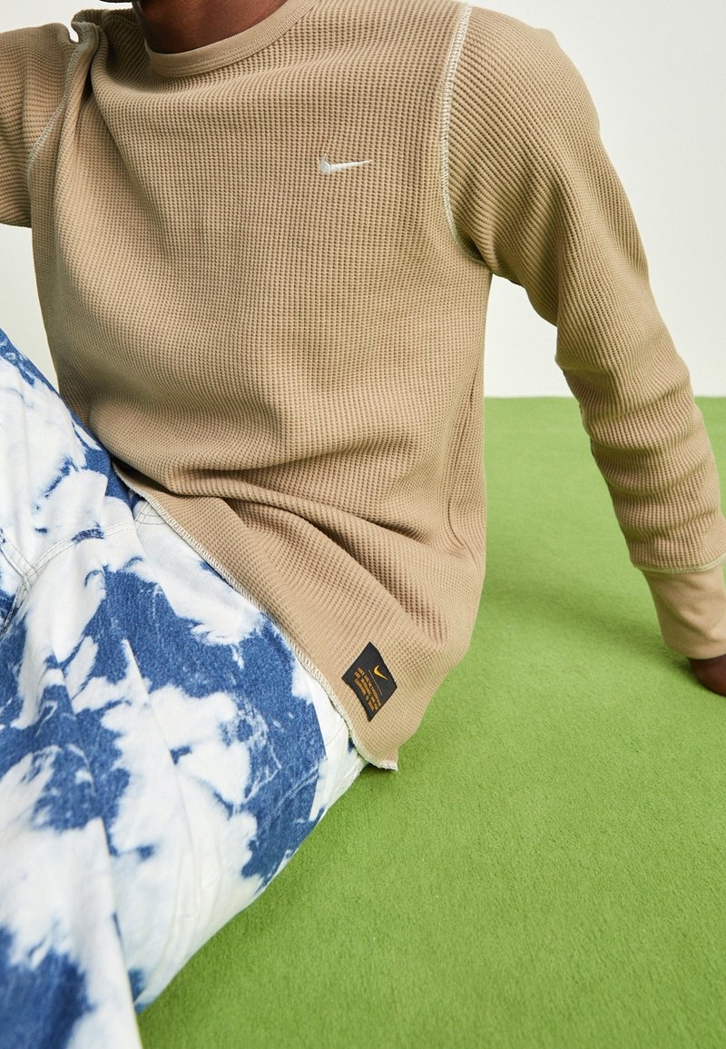 Chemise texturée beige à manches longues avec logo argenté, associée à un pantalon tie-dye bleu et blanc, sur une surface en tissu vert.