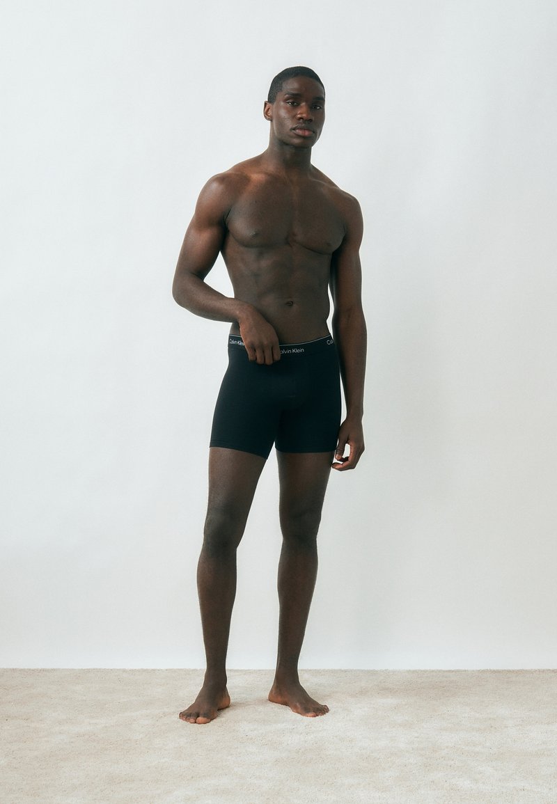 Homme musclé debout, pieds nus sur un tapis beige, portant un boxer noir Calvin Klein, devant un mur blanc uni.