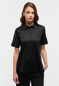 Camicia nera a maniche corte in tessuto liscio, con patta dei bottoni e colletto classico. Indossata con pantaloni a righe sottili.