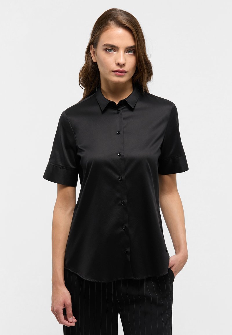 Camicia nera a maniche corte in tessuto liscio, con patta dei bottoni e colletto classico. Indossata con pantaloni a righe sottili.