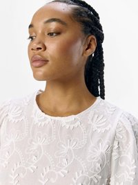Blusa bianca ricamata con motivi floreali, scollatura rotonda e maniche a palloncino. Tessuto leggero con dettagli di cucitura intricati.