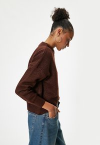 Maglione marrone strutturato con scollatura rotonda e maniche lunghe, abbinato a jeans azzurri a vita alta, mostrato in una vista di profilo.