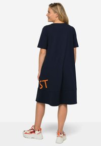 Navy blue, short-sleeve dress with a relaxed fit, featuring orange text "ST" at the hem. Soft fabric and casual design. Worn with colorful sneakers.
