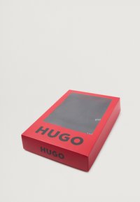 Imballaggio rettangolare rosso con parte superiore trasparente che mostra un oggetto scuro all'interno, con bold testo nero "HUGO" su due lati.
