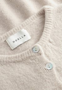 Sanftes beige gestricktes Strickjacke mit zwei Perlmuttknöpfen und einem weißen Etikett mit der Aufschrift "Noella" am Halsausschnitt.