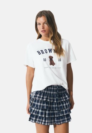 T-shirt en coton blanc avec le texte "BROWN" et un graphique d'ours ; associé à une jupe à carreaux marine superposée avec des détails en volants et une taille élastique.