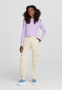Top a maniche lunghe di colore lilla con spalle scoperte, abbinato a pantaloni cargo crema con tasche laterali e polsini elasticizzati. Le sneakers azzurre leggeri completano il look.