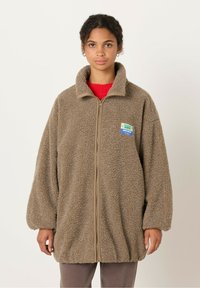 Fleece takki ruskeana, jossa on vetoketju edessä, korkea kaulus ja suuret hihat. Rinnassa on etiketti, jossa on värikäs kuvio.