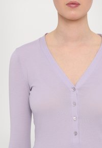 Lavendelkleurige longsleeve top met een V-hals en drie-knopen sluiting. Het materiaal is glad en aansluitend.